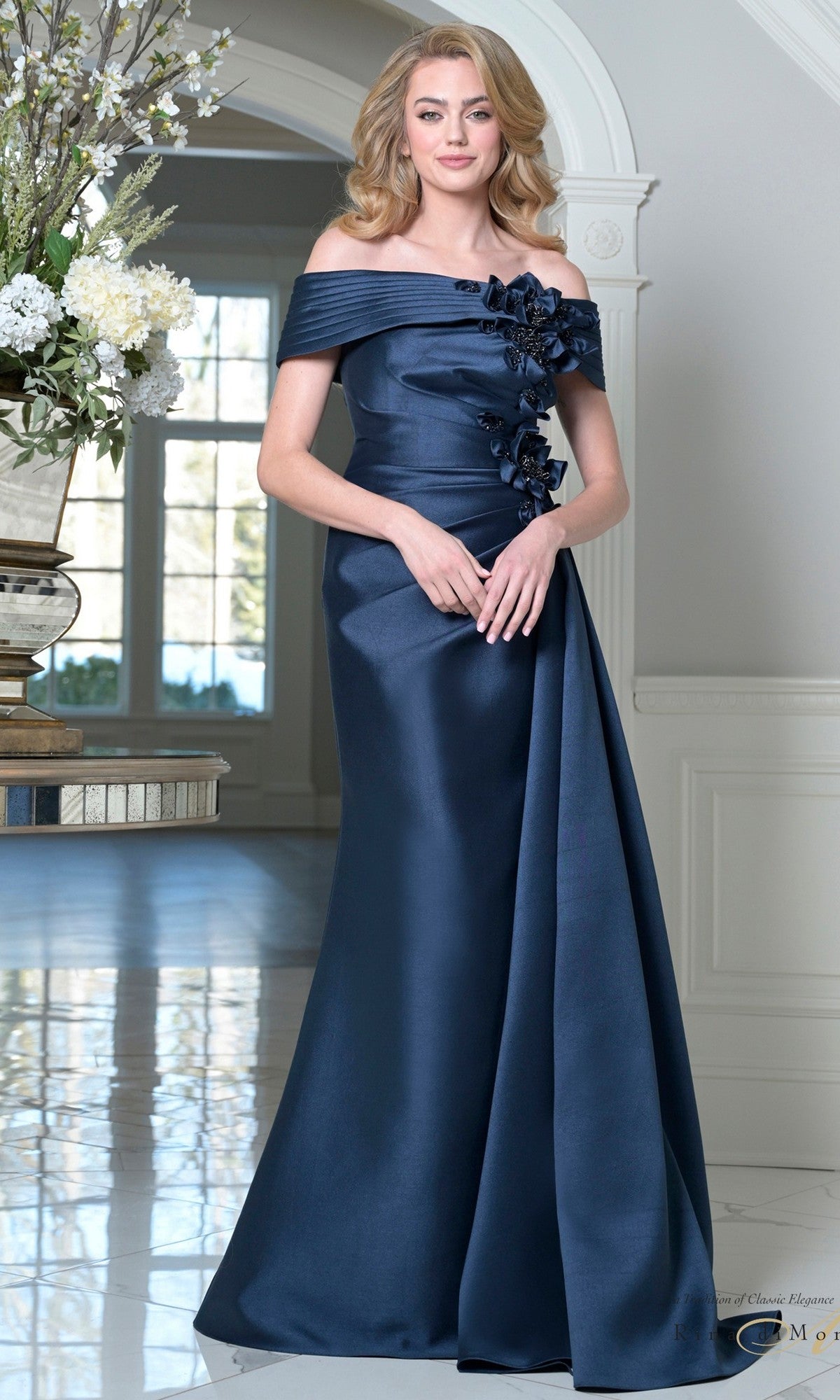 A woman stands indoors by a large window and floral display, wearing the Long Formal Dress: Rina Di Montella RD3528 in navy, featuring an elegant off-shoulder neckline and 3D floral appliques.